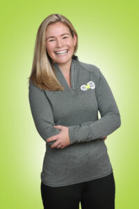 Smiling woman with long blonde hair in a gray athletic top, arms crossed, against a lime green background.