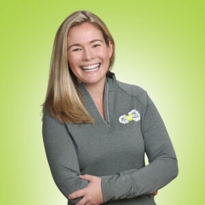 Smiling woman with long blonde hair wearing a gray zip-up athletic top, arms crossed, against a lime green background.