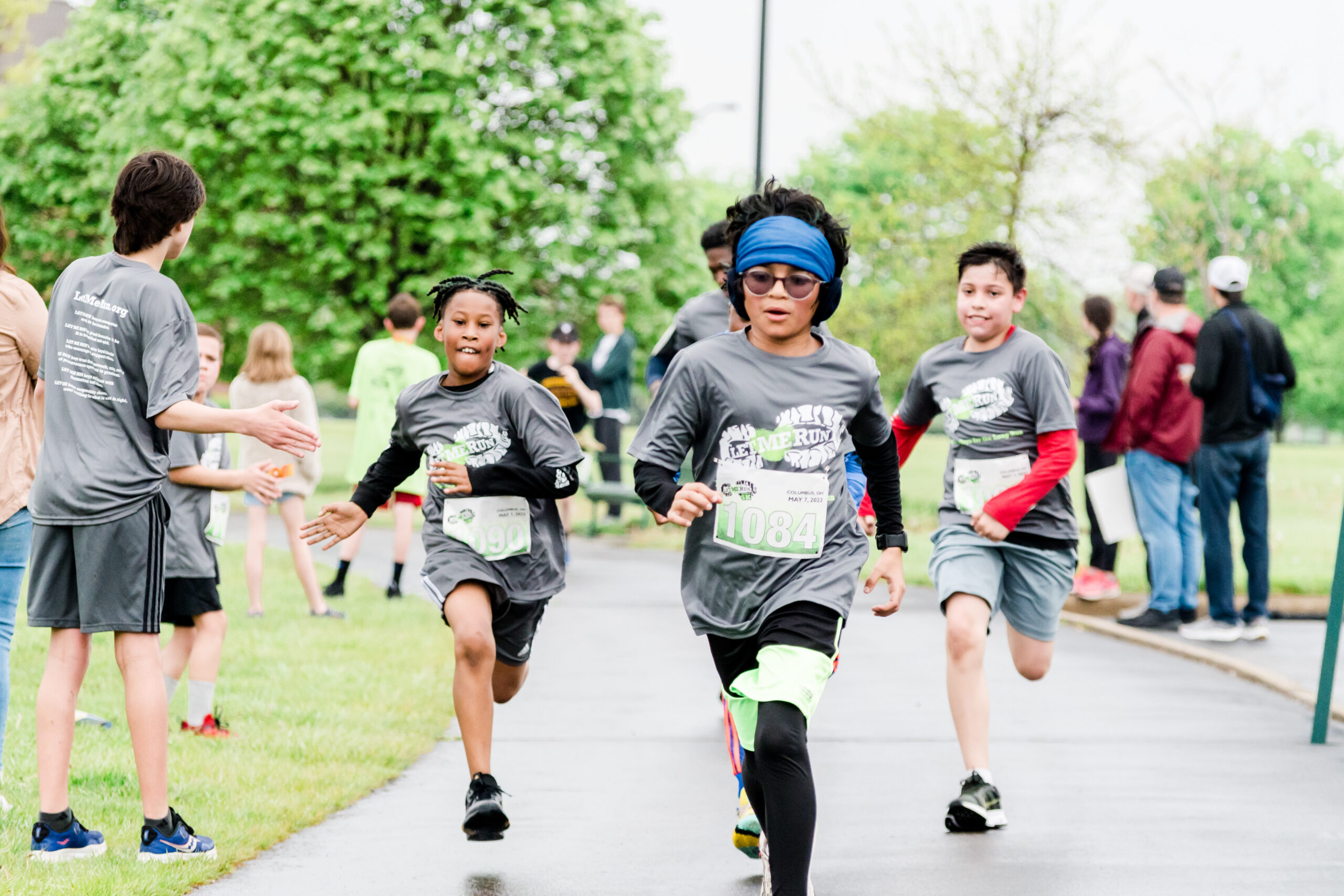 Group of children running a race on a park path, wearing matching gray shirts and numbered bibs on the front gear.