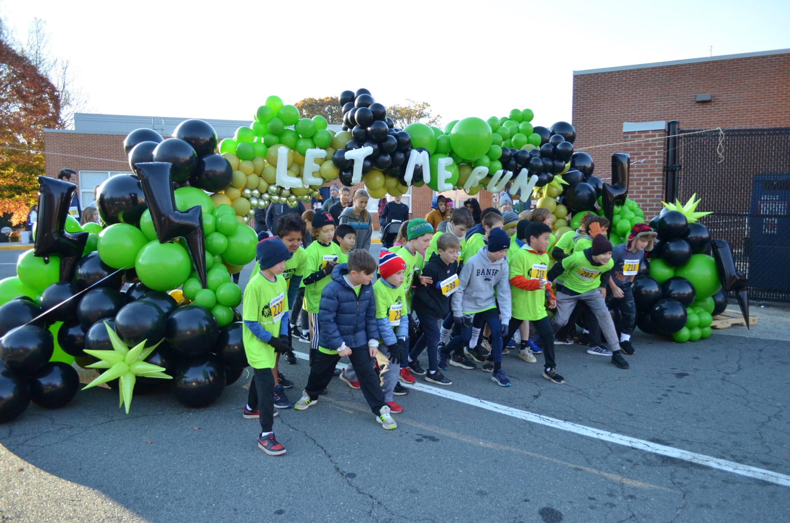Children in neon shirts start a race under a green/black balloon arch that reads 'LET ME RUN' on a sunny street.