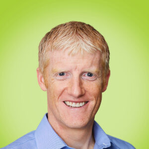 Smiling man with blonde hair wearing a light blue collared shirt against a lime green background.