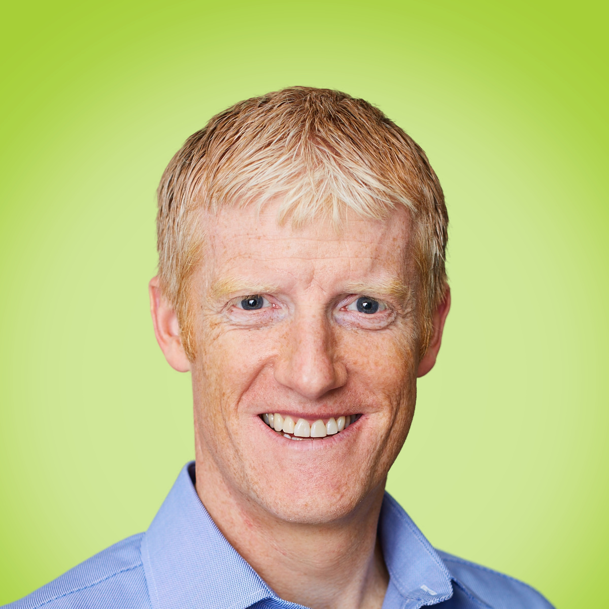 Smiling man with blonde hair wearing a light blue collared shirt against a lime green background.